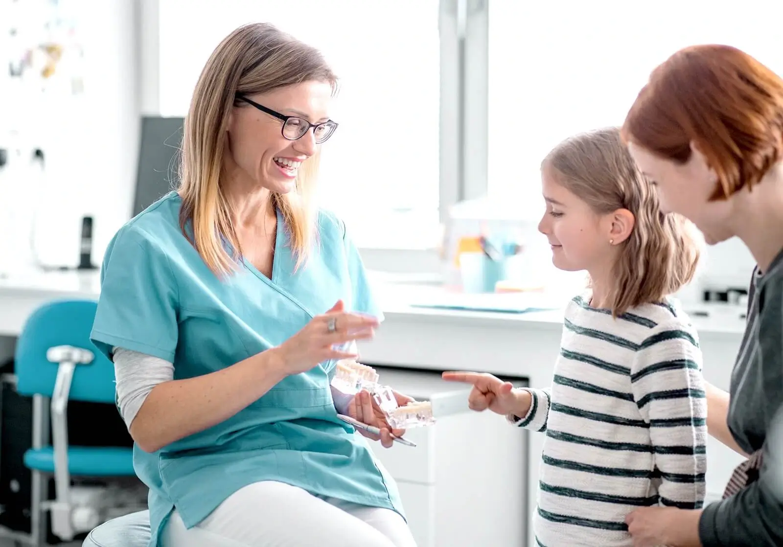 A dental hygienist speaks with a young patient and her mother in a dental clinic